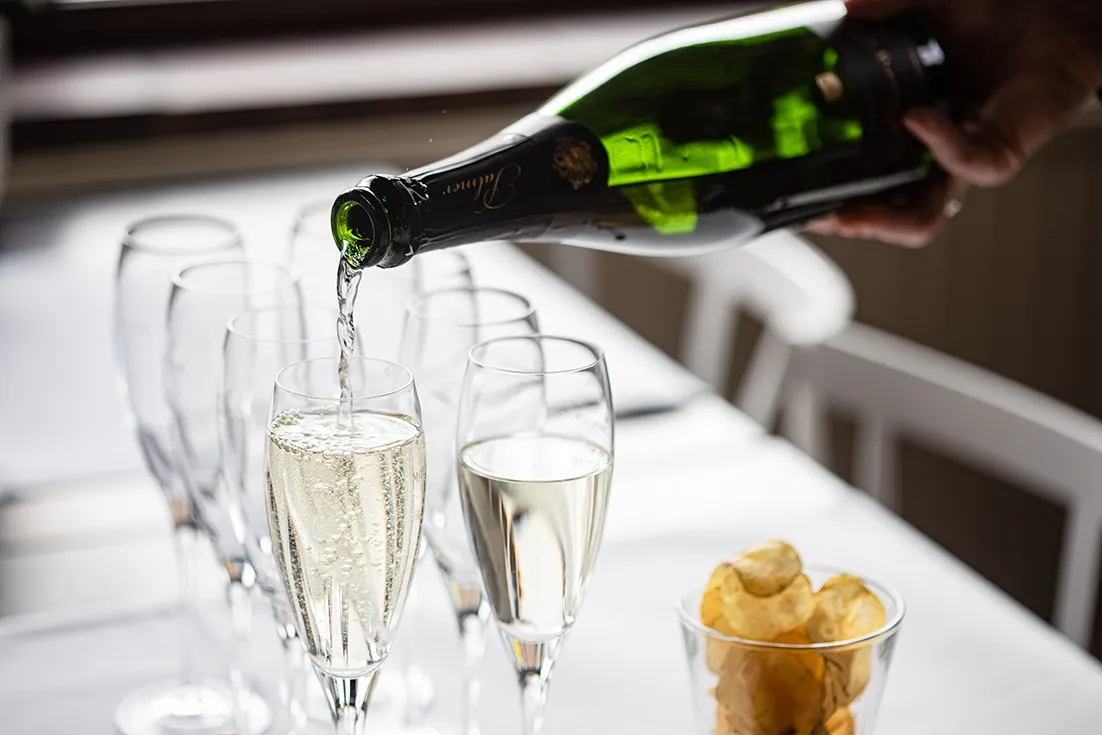 En flaska mousserande vin som häller upp i ett champagneglas, med fler glas i bakgrunden på ett vitt bord.